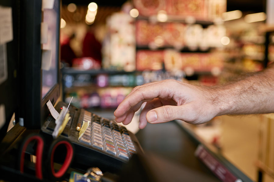 A hand over an old-style cash-register keypad with paper receipts and blurred shop shelves behind